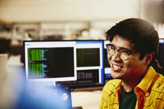 Man smiling while sitting behind his desk, computer screens displaying programming code.