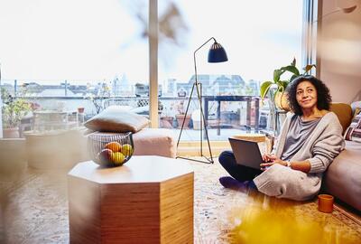 Woman leaning against a couch, sitting on the floor, with laptop on her lap