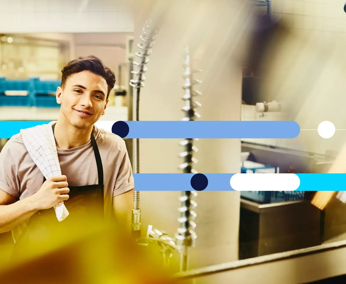 Smiling male looking straight into the camera with dishtowel on shoulder standing next to the dishwasher station in a kitchen.