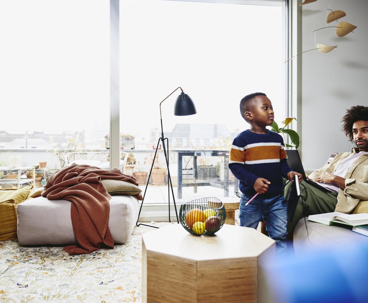 Man sitting on couch, working on laptop. Kid holding a notebook and pen.