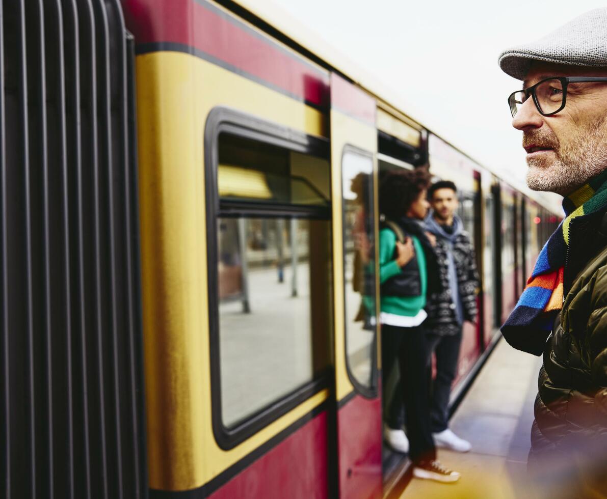 Man standing next to a train. Two people getting out of a train in the background.
