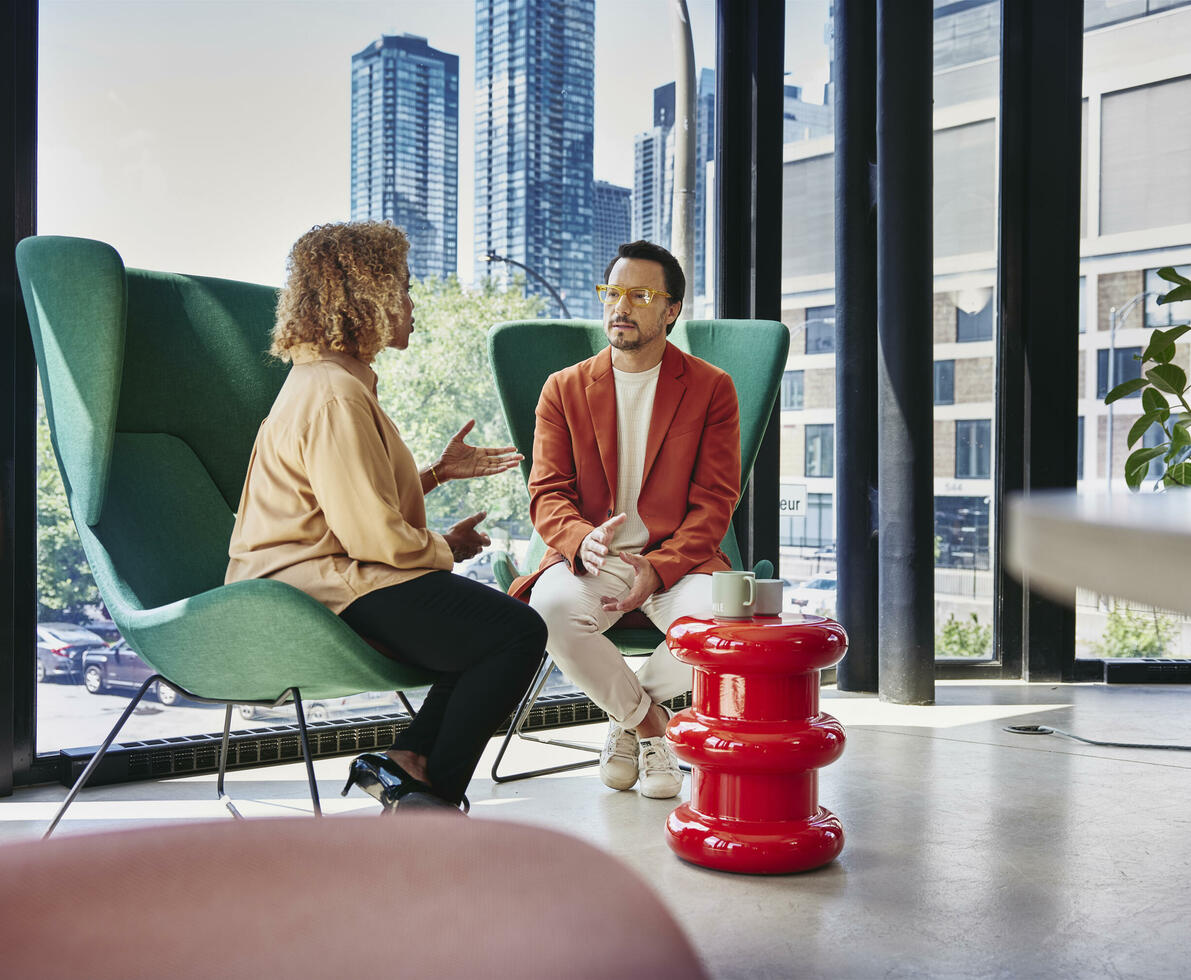 Male with glass and red blazer sitting in a green chair talking to a female sitting in a green chair