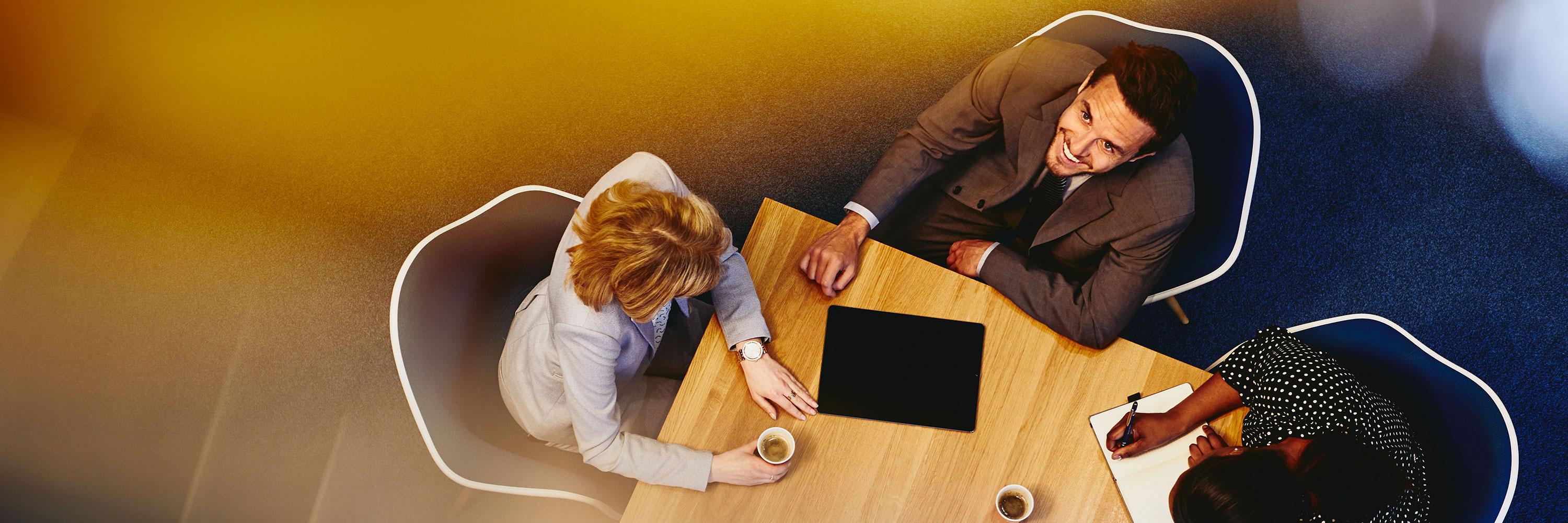 People in an office having a meeting, taking notes, coffee. Man smiling and looking up.