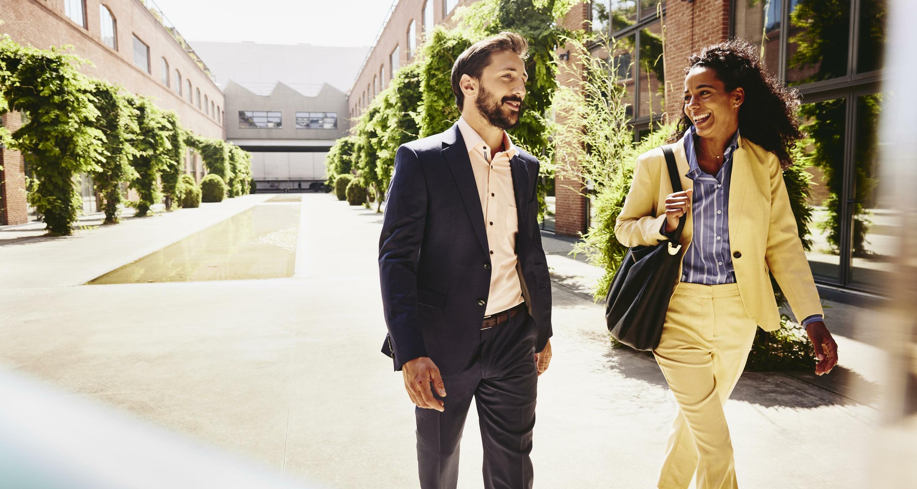 Man and woman talking and having a laugh while walking outside between office buildings.